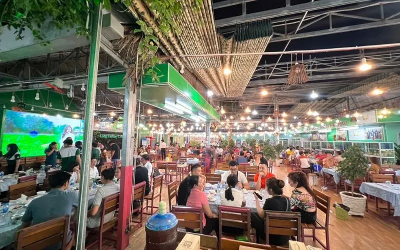 Không gian sảnh tiệc rộng lớn trong nhà được trang bị hệ thống đèn sáng sủa, tấp nập khách đoàn lữ hành tại Làng Nướng Châu Đốc